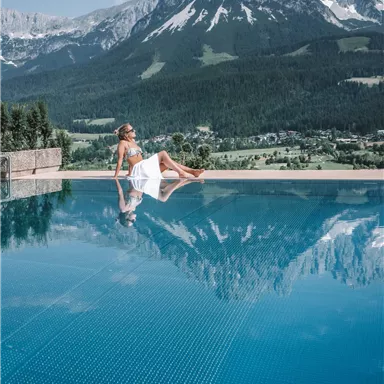 A woman relaxes by the edge of a pool with a view of impressive mountains. The sky is clear and the landscape is green and inviting.