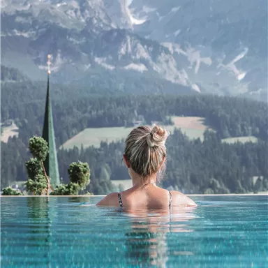 A woman is standing in the water of an infinity pool, looking at the mountains. In the background, green meadows and a church tower are visible.