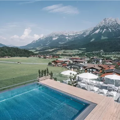 A beautiful view from a pool overlooking a picturesque landscape with mountains and a small village. The surroundings are green and inviting, ideal for relaxation and nature experiences.