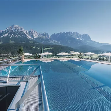 An infinity pool overlooking impressive mountains and a clear blue sky. Sun loungers are ready for relaxation.