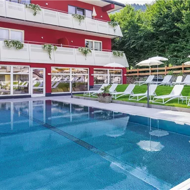 A beautiful outdoor pool with loungers and umbrellas. In the background stands a red building surrounded by trees.