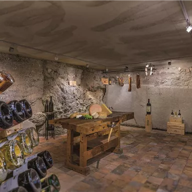 A cozy wine cellar with natural stone walls and warm light. On the shelves, bottles and cheese can be seen, and in the center stands a large wooden table.