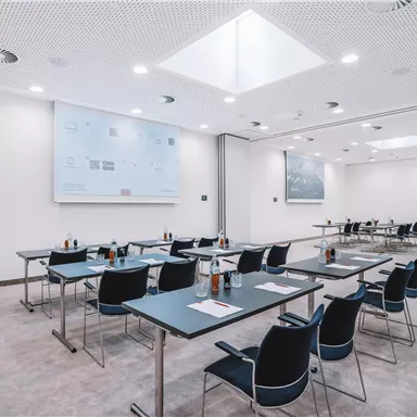 A modern seminar room with tables and chairs.  
There is a screen on the wall and the lighting is bright.