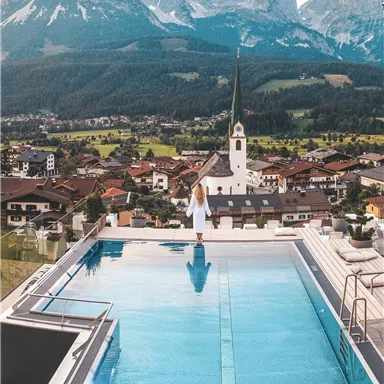 A breathtaking view of the mountains and a village with a church. A person stands at the edge of an infinity pool and enjoys the view.