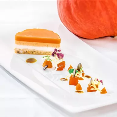 An artistically arranged dessert with layers of pumpkin and a decoration of colorful vegetables. In the background lies a large pumpkin.