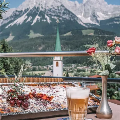 A beautifully set table with a cake and a glass of beer. In the background, majestic mountains and a church steeple can be seen.