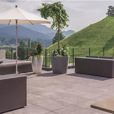 A modern terrace with comfortable lounge furniture and sun umbrellas. In the background, green meadows and mountains can be seen.