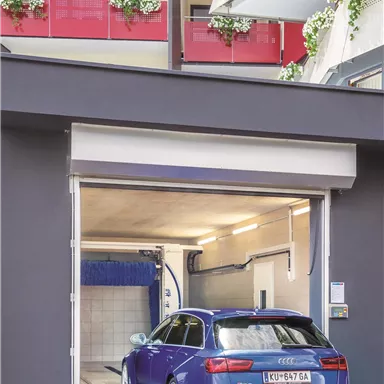 A blue Audi is in front of an open garage door. The background shows a modern building complex with balconies.