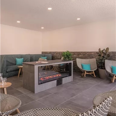 A modern relaxation room with comfortable seating and a central fireplace. The walls are painted in neutral colors, complemented by green plants.