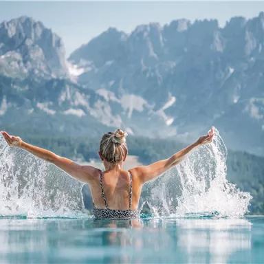 A woman is sitting in an infinity pool and splashing water. Majestic mountains and a picturesque landscape are visible in the background.