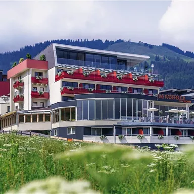 A modern hotel complex amidst a green landscape. In the background, hills and a cloudy sky can be seen.