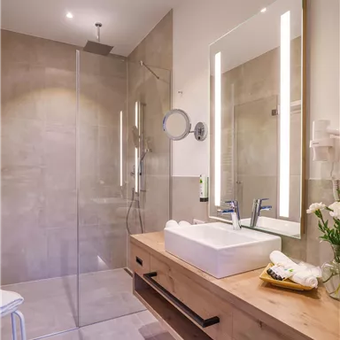 A modern bathroom with a clear shower and an elegant wooden sink. The room has bright lighting and a simple, elegant design.