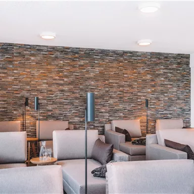 A modern lounge area with comfortable, gray sofas and atmospheric lighting. A stone wall and plants give the space a relaxed atmosphere.