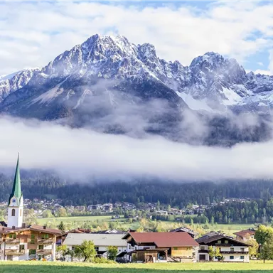 Eine malerische Berglandschaft mit schneebedeckten Gipfeln und einem klaren Himmel. Im Vordergrund befindet sich ein kleines Dorf mit einer Kirche und grünen Wiesen.