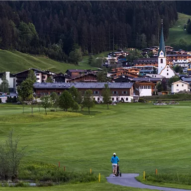 A picturesque village landscape with green meadows and traditional architecture. In the background, a church tower rises between the houses.