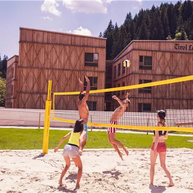 Ein Sandvolleyballspiel im Freien mit mehreren Spielern. Im Hintergrund sind moderne Holzhäuser und grüne Wälder zu sehen.
