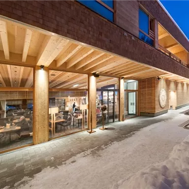 Ein modernes Gebäude mit großen Fenstern und Holzfassade, umgeben von Schnee. Im Inneren sind Menschen sichtbar, die gemütlich sitzen und sich unterhalten.