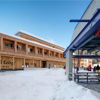 A modern wooden building in the snow with the inscription "Tire Lodge". In the foreground, a ski lift and some visitors can be seen.