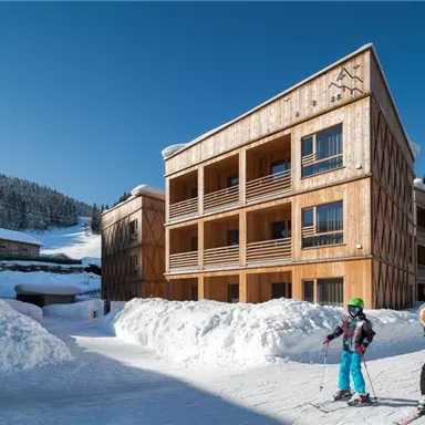 A wooden facade of the building in the snow, surrounded by snow-covered mountains. Two skiers are standing on the path and enjoying the surroundings.