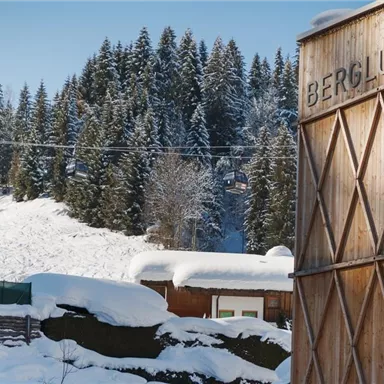 A snowy mountain landscape with dense coniferous trees and a ski lift in the background. In the foreground, there is a wooden building with the inscription "BERGLUFT".