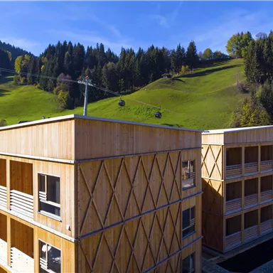 Moderne Holzbauten in einer hügeligen Landschaft. Im Hintergrund sind grüne Wiesen und Bäume zu sehen.