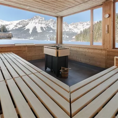 A cozy sauna area with wooden benches and a fireplace. In the background, snow-covered mountains stretch under a blue sky.