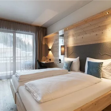A modern bedroom with two beds and stylish wooden decor. Large windows provide plenty of daylight and a view outside.