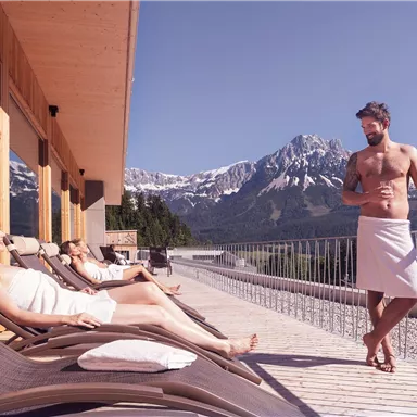 Eine entspannte Wellness-Szene auf einer Terrasse in den Bergen. Menschen in Bademänteln genießen die Aussicht auf die Alpen.