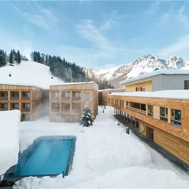 A modern hotel building in a snow-covered landscape. In the foreground, there is a heated pool, and in the background, snow-capped mountains can be seen.