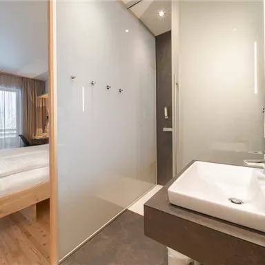 A modern hotel room with a clean and minimalist design. On the left, you can see a comfortable bed, and on the right, a bathroom with an elegant sink.