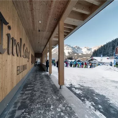 A modern entrance area with wood paneling in a snow-covered environment. In the background, people can be seen preparing for a ski trip.