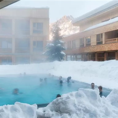 A pool in the middle of snow with several people relaxing in the water. In the background, the wooden buildings of an accommodation can be seen.