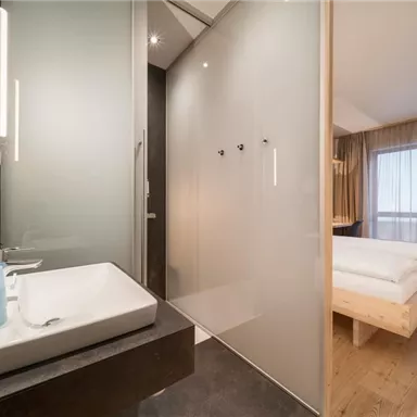 A modern bathroom with a sink and a glass door shower cabin. In the background, there is a bright bedroom with a large window and a wooden bed.