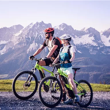 Ein paar Fahrradfahrer genießen die Aussicht auf eine beeindruckende Berglandschaft. Die Umgebung ist grün und die Berge sind schneebedeckt.