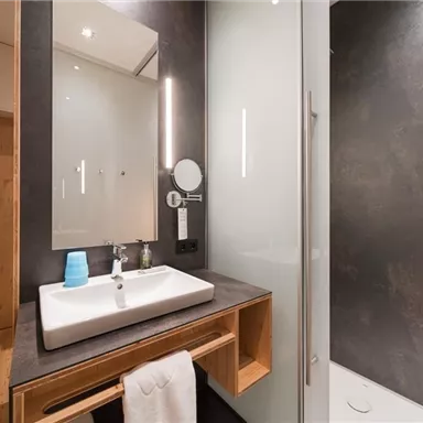 A modern bathroom with an elegant sink unit and a large mirror. The walls are dark and the shower is enclosed with glass.