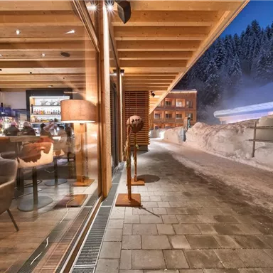 A modern restaurant with wooden decor and a cozy atmosphere. Outside, snow lies on the ground and the surroundings are surrounded by wooded mountains.