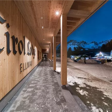 A modern wooden entrance with the inscription "Tirol" and a view of snow-covered mountains. It is dusk, and the surroundings are calm and inviting.