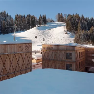 A modern wooden building in the mountains, surrounded by snow-covered hills. In the background, ski lifts and a snowy slope are visible.