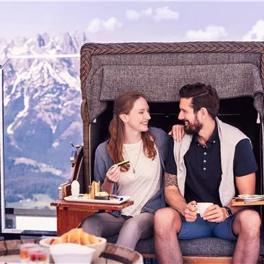 Ein Paar sitzt in einem gemütlichen Strandkorb mit Blick auf die Berge. Sie lächeln sich an und genießen ihre Zeit zusammen.