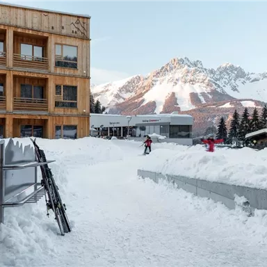 A picturesque winter resort with snow-covered mountains in the background. In the foreground, there is ski equipment and people walking in the snow.