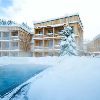 A modern hotel in a snowy landscape. The exterior features a pool, surrounded by snow and wooden architecture.