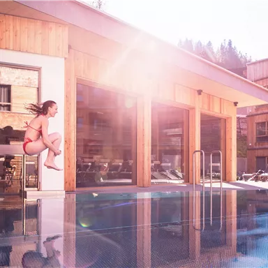 Ein Mädchen springt in einen Pool vor einem modernen Holzgebäude. Die Sonne scheint und es ist ein klarer, schöner Tag.