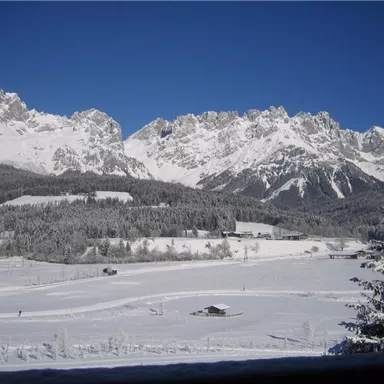 A snowy mountain landscape under a clear blue sky. In the foreground, snow-covered fields and a few small buildings can be seen.