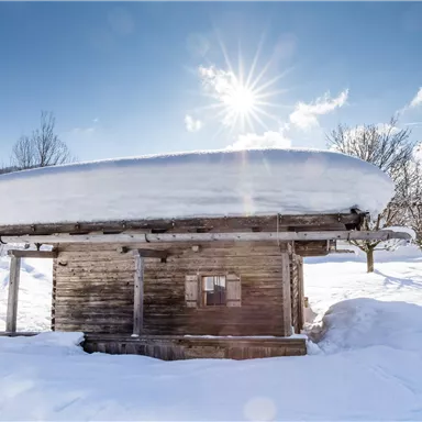 A cozy wooden cabin with a snow-covered roof. The sun is shining clearly in the blue sky and the snow blankets the landscape.