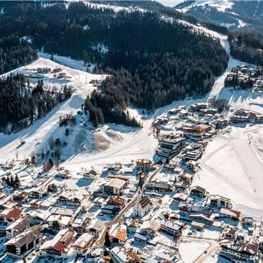 A picturesque winter landscape with snow-covered mountains and a small village. The buildings are surrounded by snow, and the slopes are prepared for skiing.