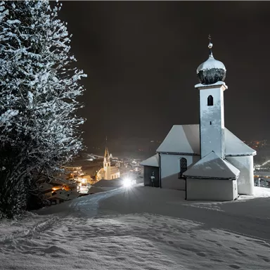 Eine schneebedeckte Landschaft bei Nacht mit einer kleinen Kirche und einem hohen Kirchturm. Im Hintergrund leuchten die Lichter eines kleinen Dorfes.