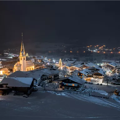 Eine verschneite Stadt bei Nacht mit warmen Lichtern. Im Hintergrund ist eine Kirche mit einem hohen Turm zu sehen.