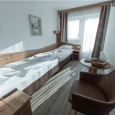 Ein einfaches Hotelzimmer mit zwei Einzelbetten und einem bequemen Sessel. Große Fenster lassen viel Licht herein und bieten einen Blick nach draußen.