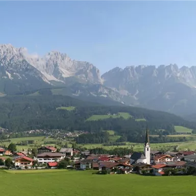 Eine malerische Berglandschaft mit beeindruckenden Gipfeln im Hintergrund. Im Vordergrund liegt ein kleines Dorf in einer grünen Talebene.