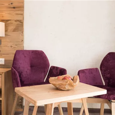 Ein gemütlicher Wohnbereich mit zwei lila Stühlen und einem Holztisch. Auf dem Tisch liegt eine Schale mit Äpfeln.